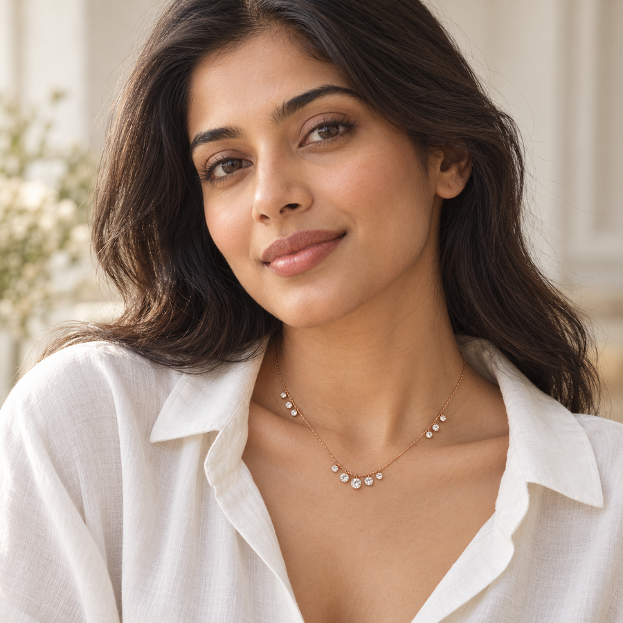 A woman with dark hair wearing a delicate gold necklace with small white stones, styled with a white linen shirt against a softly blurred background.