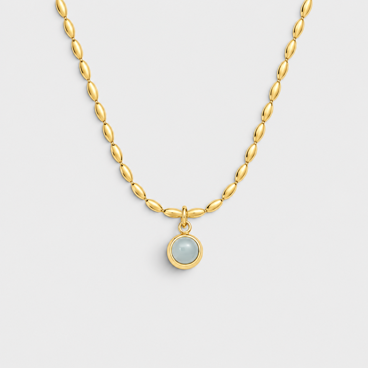 A gold chain necklace with a small oval pendant featuring a pale blue-gray stone in a gold bezel setting, shown on a light gray background.