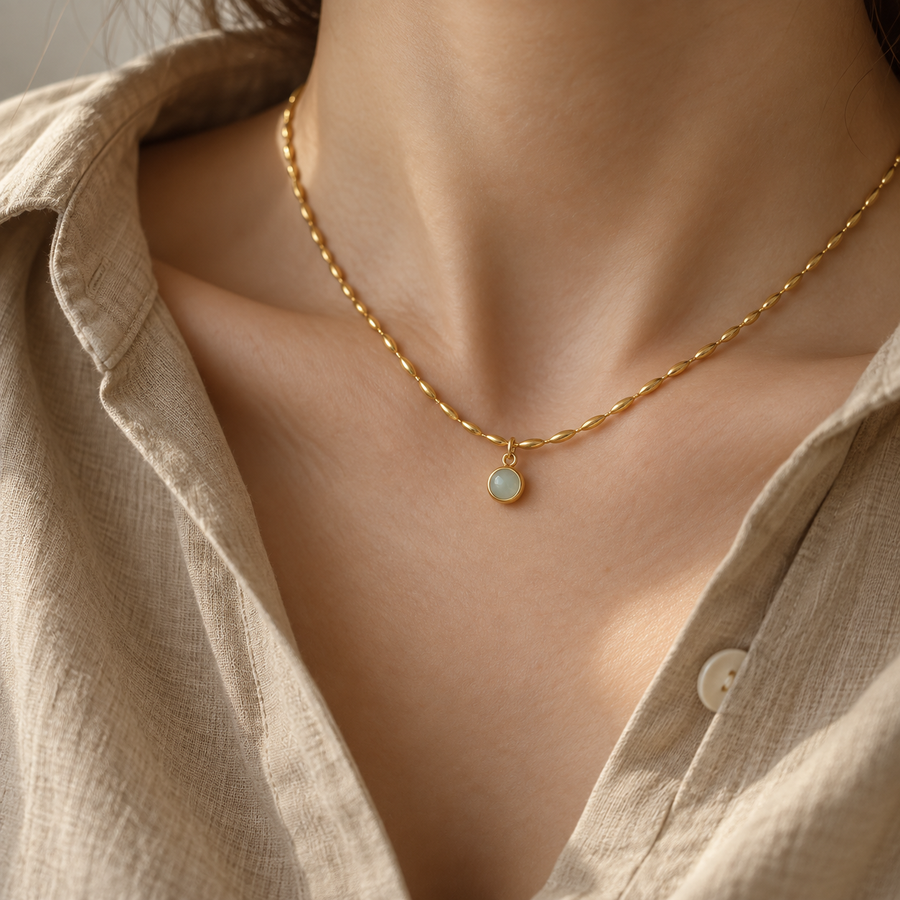 A close-up of a person's neck and chest wearing a delicate gold chain necklace with a small round light blue stone pendant, paired with a beige linen shirt.