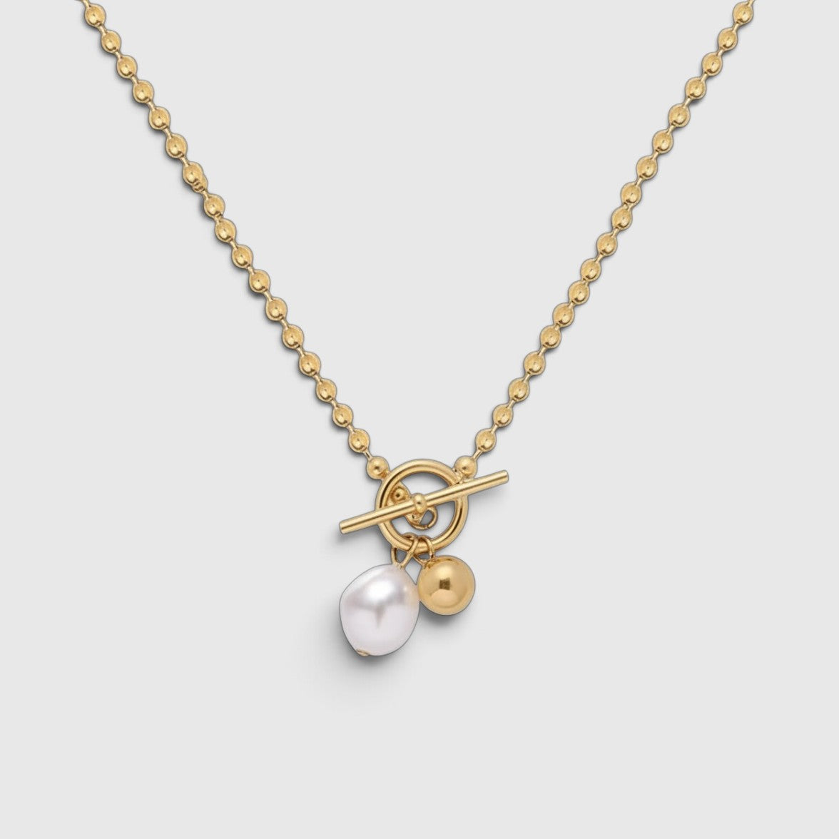 A gold necklace with a beaded chain, toggle clasp, and a dangling white pearl and gold bead charm, shown on a light gray background.