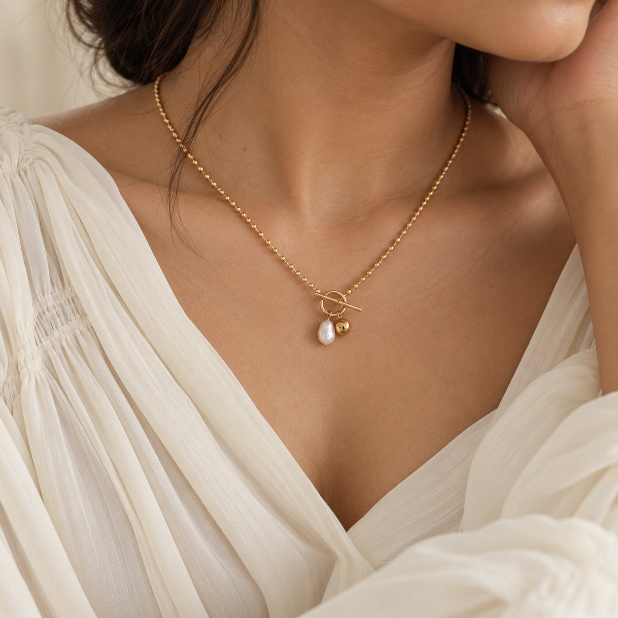 A close-up lifestyle shot of a model wearing a delicate gold necklace with a toggle clasp, a teardrop-shaped white pearl, and a small gold bead accent, paired with a cream-colored blouse.