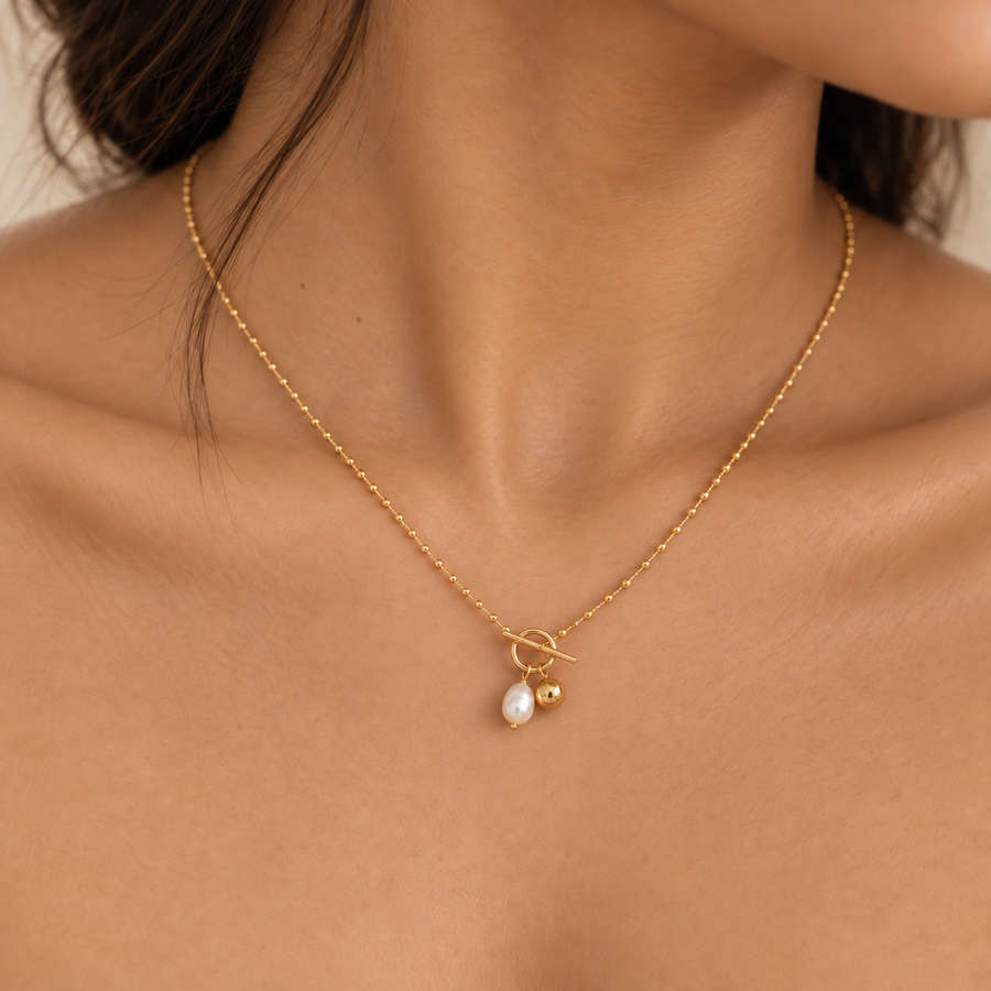 A close-up of a model's neck and upper chest wearing a delicate gold chain necklace with a toggle clasp, a small gold bead, and a teardrop-shaped white pearl pendant.