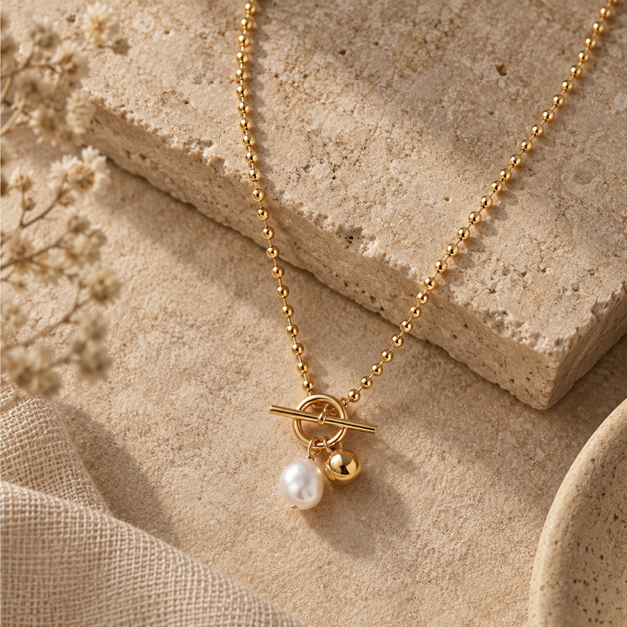 A gold-tone ball chain necklace with a toggle clasp and a dangling pearl charm, styled on a textured beige stone and linen surface with dried flowers.