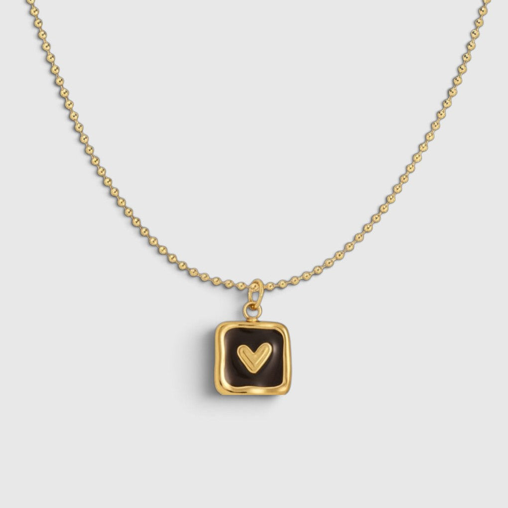 A gold ball-chain necklace with a square pendant featuring a dark brown enamel background and a gold heart in the center, displayed on a light gray background.