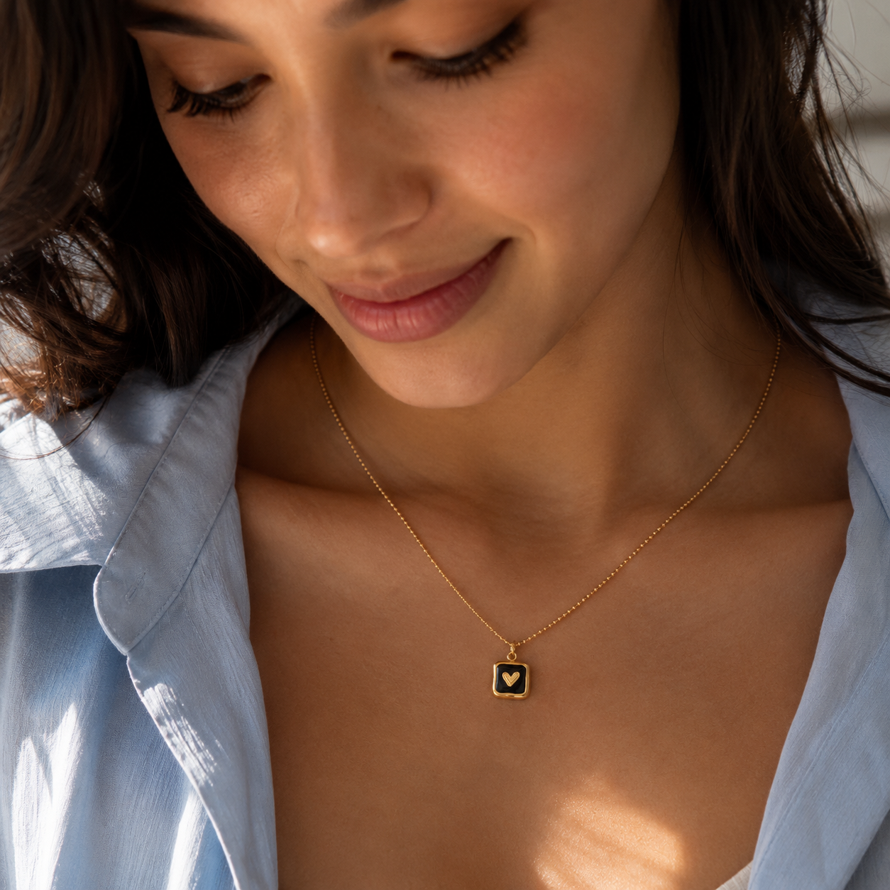 A close-up lifestyle shot of a woman wearing a delicate gold chain necklace with a square pendant featuring a black background and a small gold heart.
