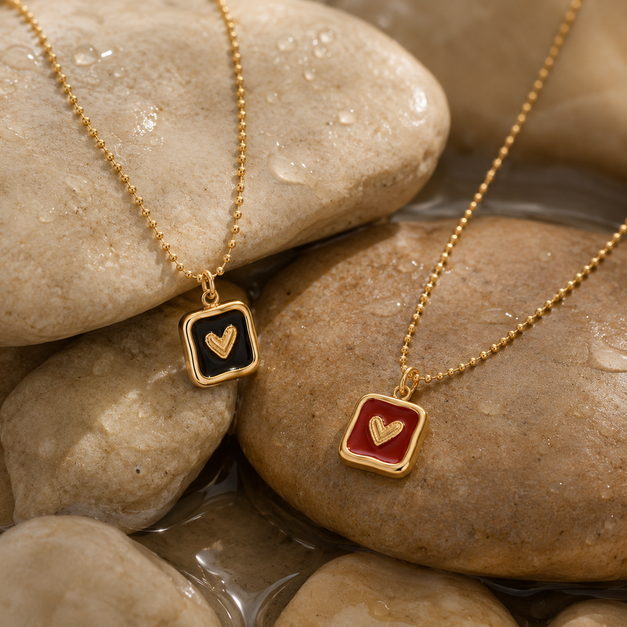 Two gold ball-chain necklaces with square pendant frames are displayed on wet stones. One pendant has a black background with a gold heart, and the other has a red background with a gold heart.