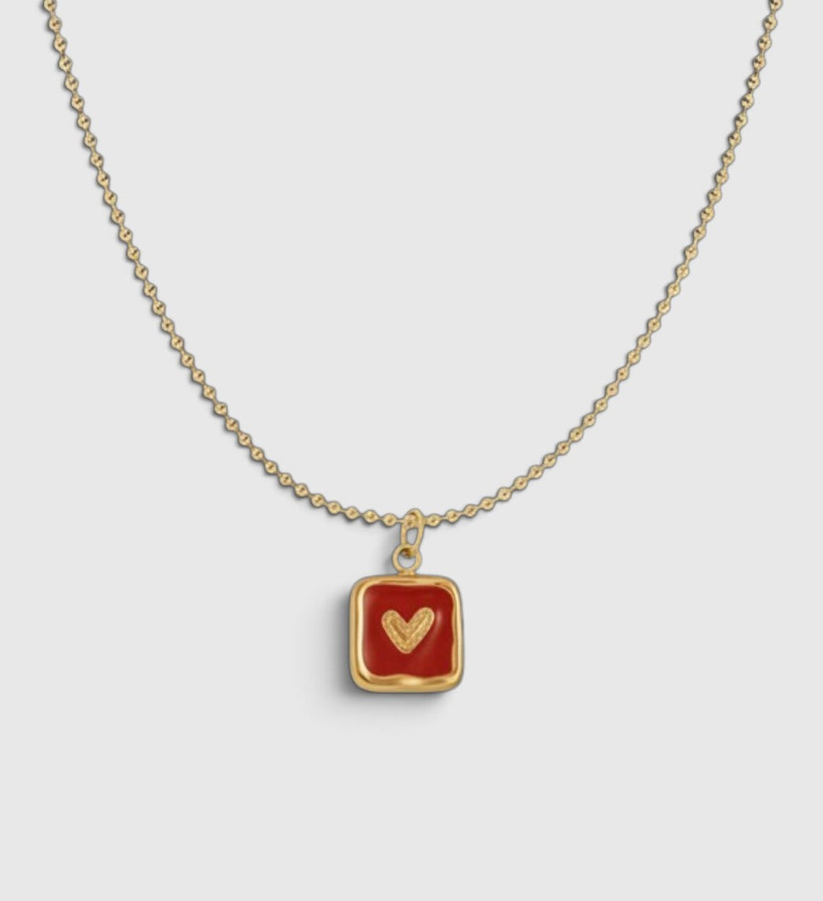 A gold chain necklace with a square pendant featuring a deep red enamel background and a textured gold heart in the center, displayed on a light gray background.