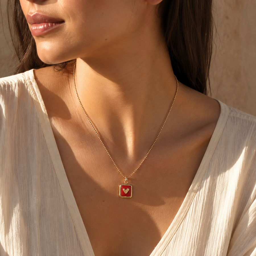 A close-up lifestyle shot of a model wearing a delicate gold chain necklace with a square pendant featuring a red background and a small white heart.