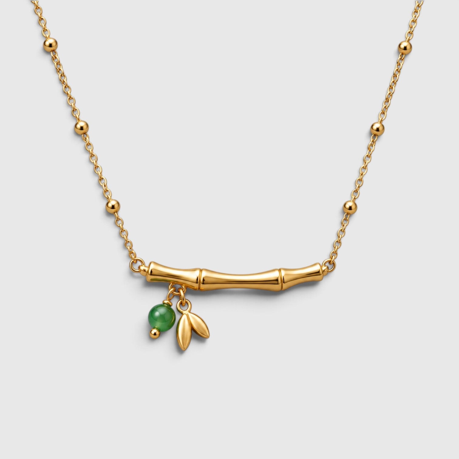 A gold chain necklace featuring a horizontal bamboo bar pendant with two small leaf charms and a dangling green bead, shown on a light gray background.