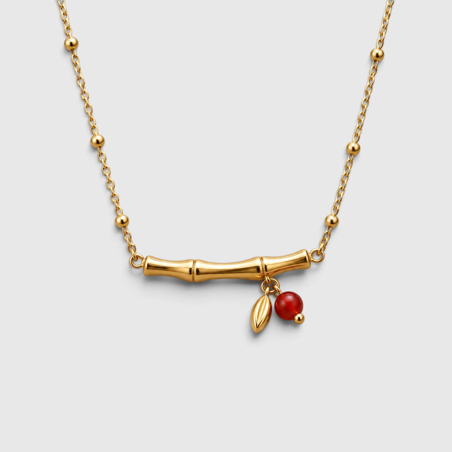 A gold chain necklace featuring a horizontal bamboo branch pendant with two small dangling charms: a red berry and a gold bead.