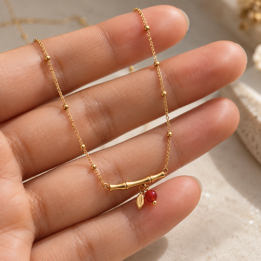 A delicate gold chain necklace with a small bamboo-like bar and a tiny red bead charm, displayed on a person's hand against a soft beige background.
