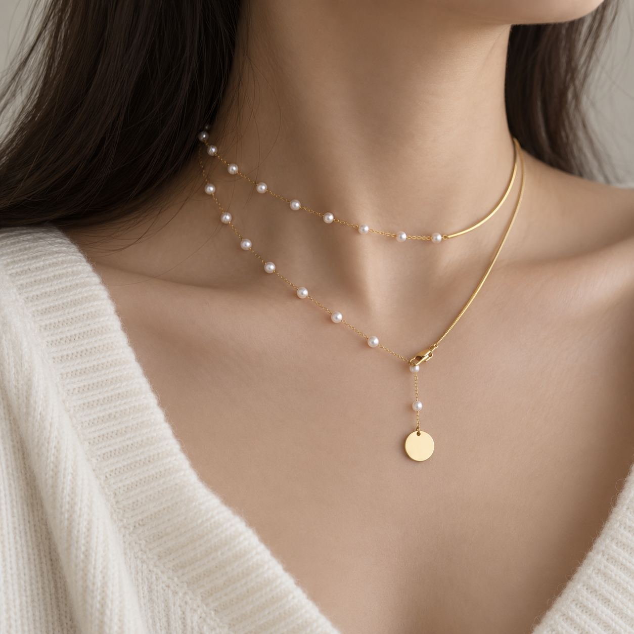 A close-up of a model wearing a delicate gold necklace with small white pearls and a round gold disc pendant, styled with a thin gold chain choker.