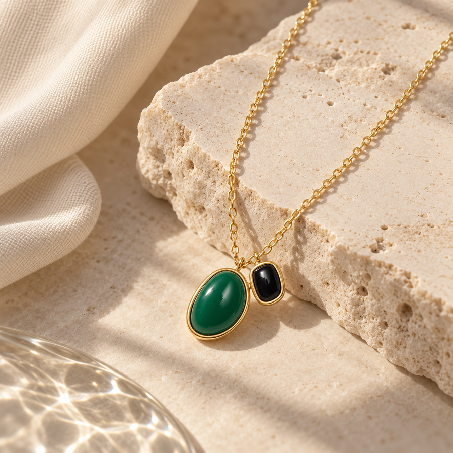 Gold chain necklace with an oval green stone pendant and a smaller black stone pendant, displayed on a textured beige stone slab with draped fabric.