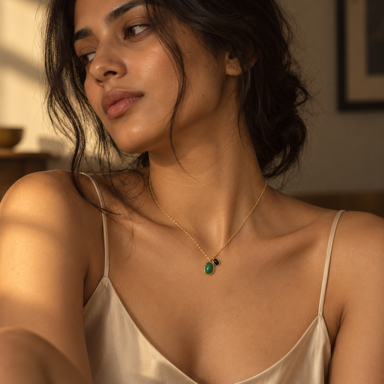 A woman wearing a delicate gold chain necklace with a small green teardrop pendant and a tiny black accent bead, styled with a cream satin camisole in warm indoor lighting.