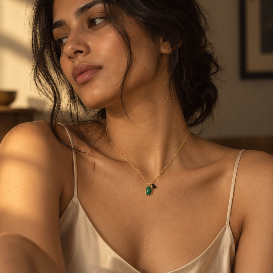 A woman wearing a delicate gold chain necklace with a small green teardrop pendant and a tiny black accent bead, styled with a cream satin camisole in warm indoor lighting.