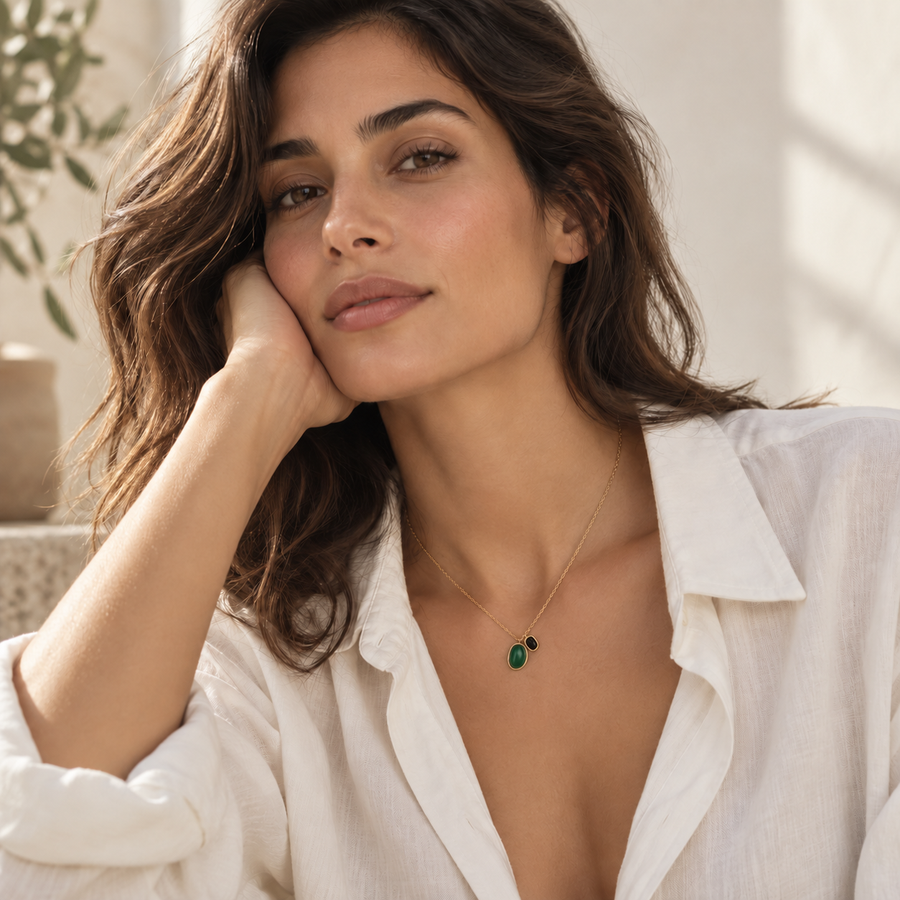 A woman with wavy brown hair resting her head on her hand, wearing a delicate gold chain necklace with a small green teardrop pendant and a tiny black accent bead, paired with a white linen shirt.