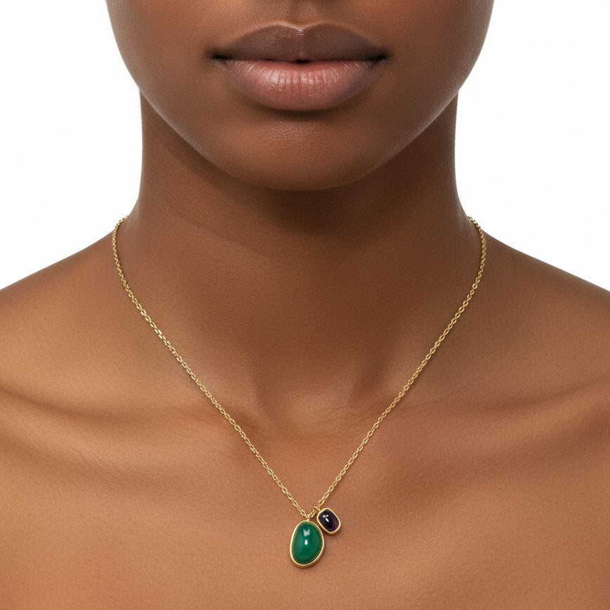 A gold chain necklace with a teardrop-shaped green pendant and a small dark round accent, shown worn on a model's neck against a white background.