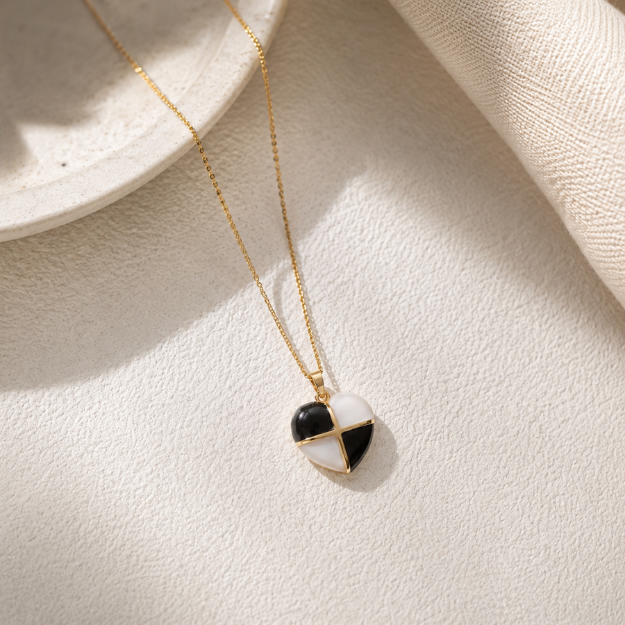 A gold chain necklace with a heart-shaped pendant featuring a black and white split design, styled on a textured cream surface with soft shadows.
