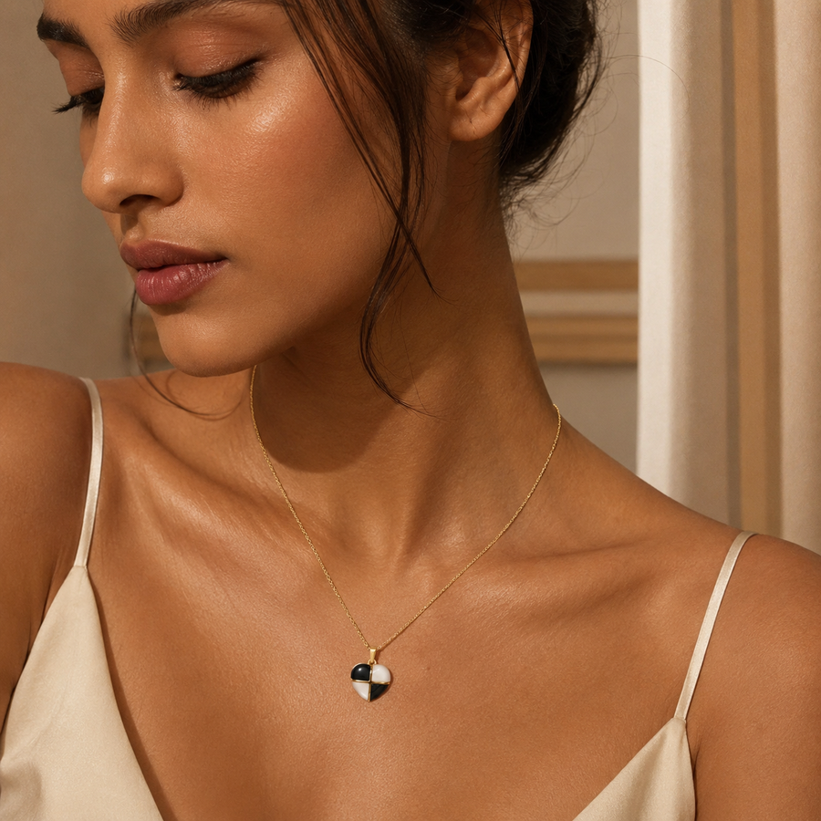 A close-up lifestyle shot of a woman wearing a delicate gold necklace with a small heart-shaped pendant featuring black and white enamel details.