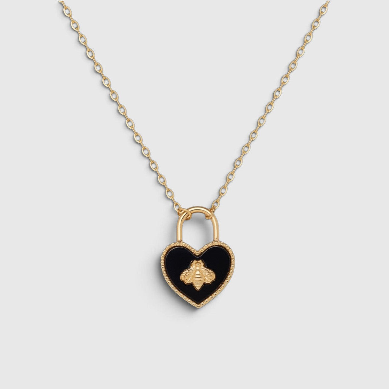 A gold heart-shaped padlock pendant necklace with a black enamel center and a gold bee motif, suspended on a delicate gold chain against a light gray background.