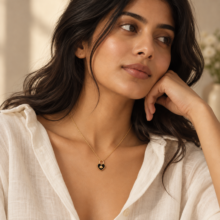 A woman wearing a delicate gold chain necklace with a small black heart pendant, styled with a white linen top in a warm, softly lit indoor setting.