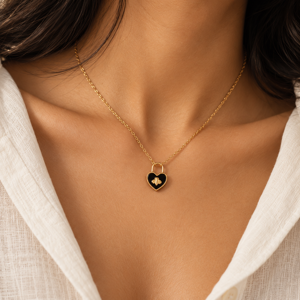 A close-up of a model wearing a delicate gold chain necklace with a small heart-shaped pendant. The pendant features a glossy black enamel center with a small gold monogram logo, set against a model wearing a white linen top.