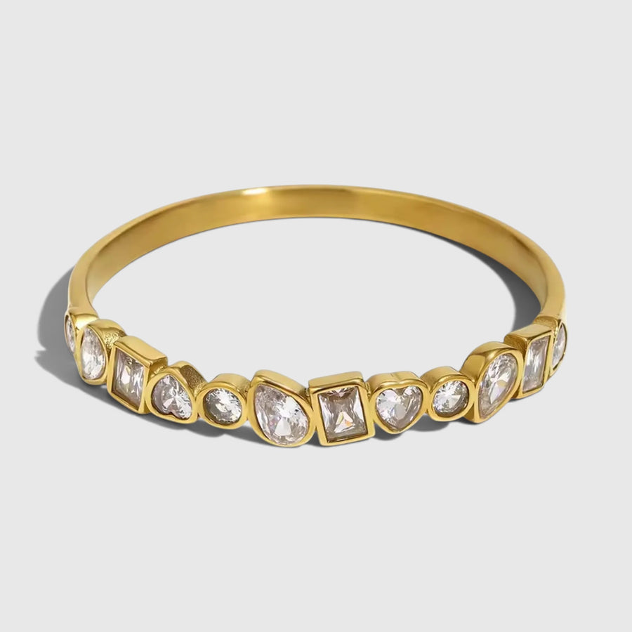 A gold bangle bracelet featuring a row of clear teardrop and square-cut gemstones in bezel settings, shown on a light gray background.
